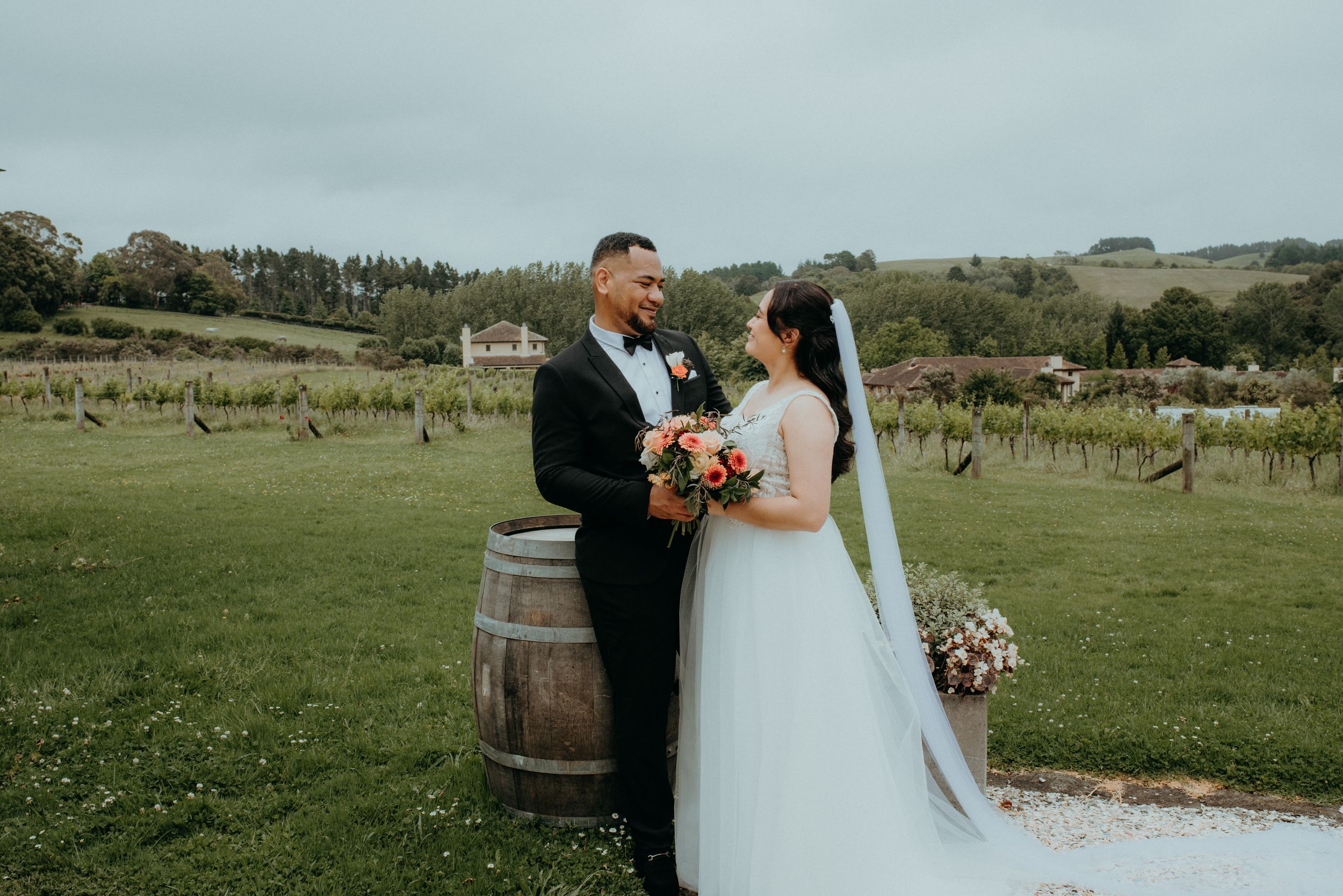 Couple in wedding attire stand together smiling by a barrel with vineyard rows behind them.