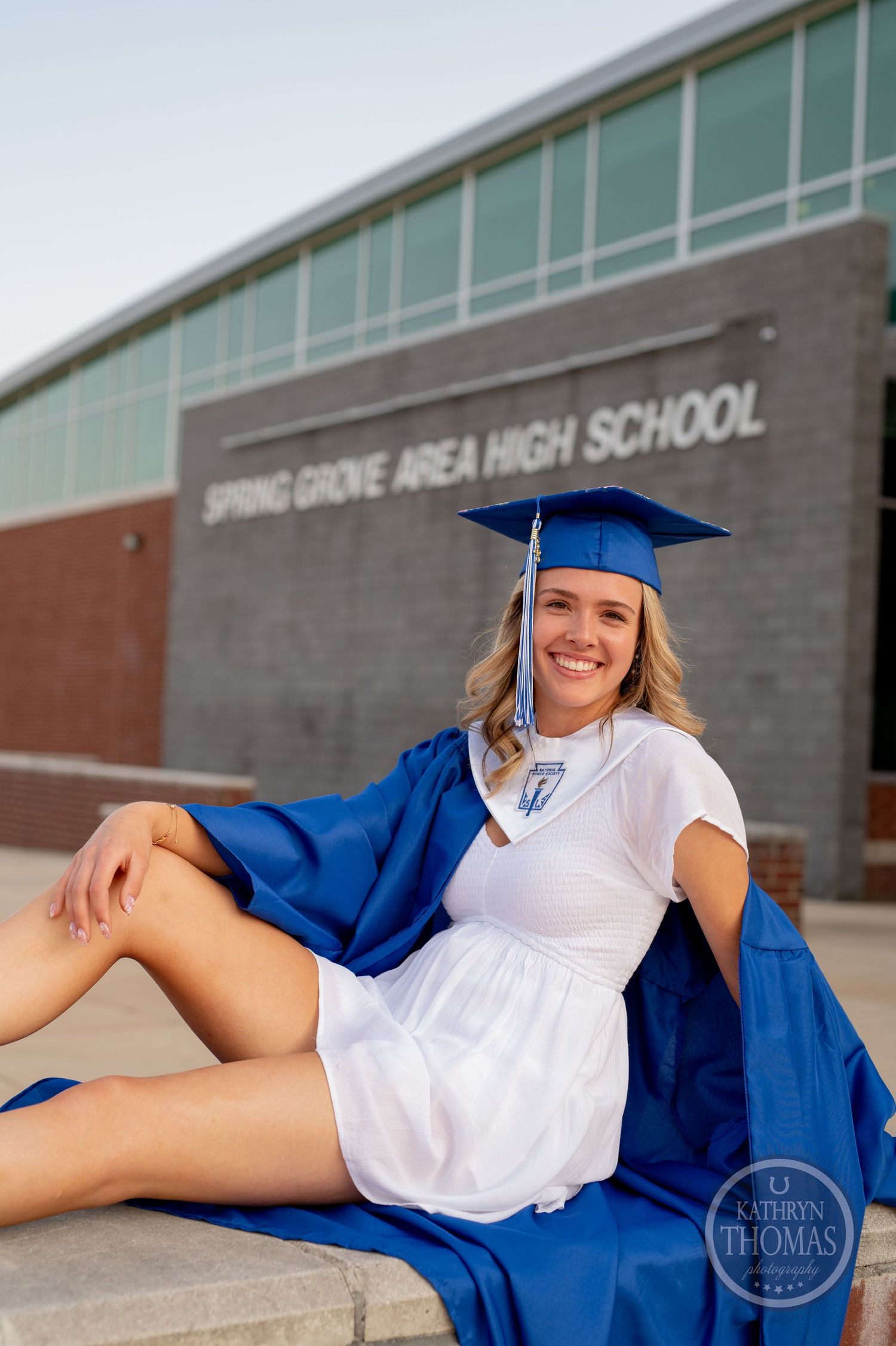 Graduate in blue cap and gown lounging on steps outside Spring Grove Area High School.