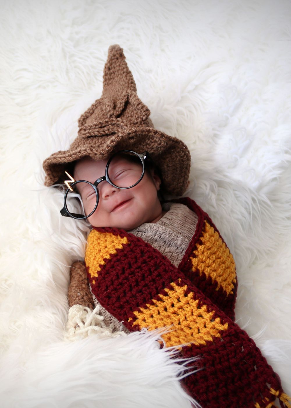 Baby in Harry Potter themed knit scarf and hat with round glasses on white fur backdrop.
