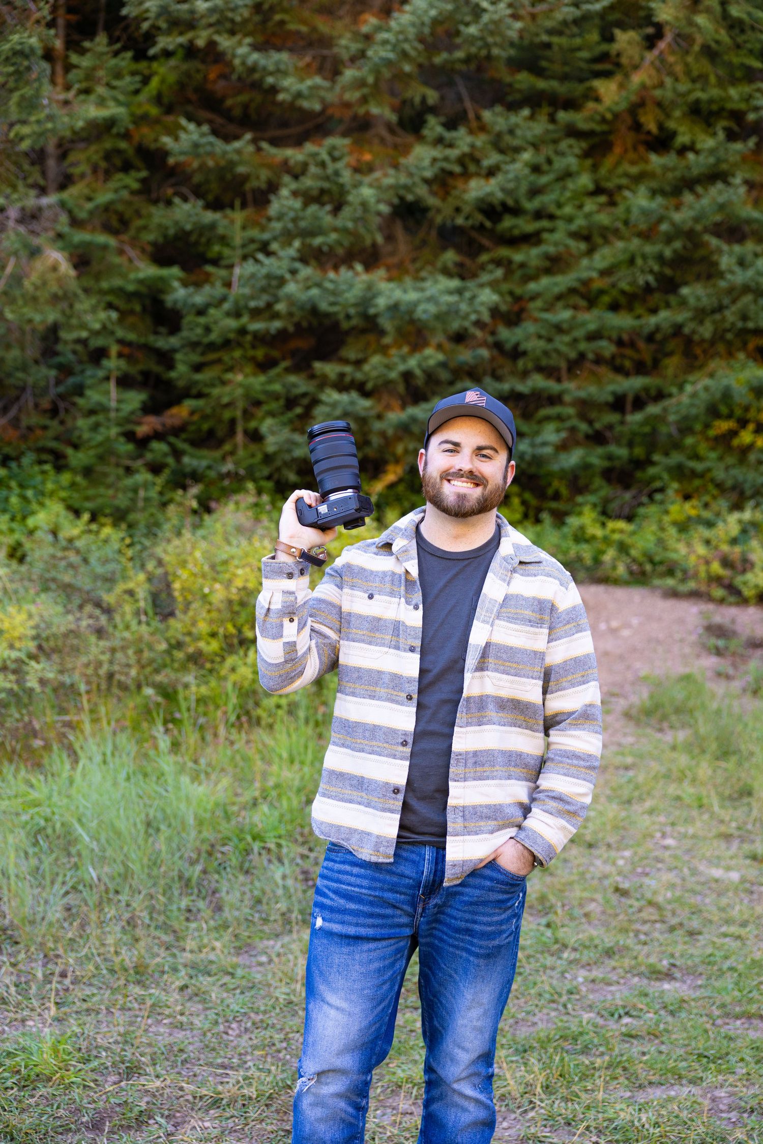 Person in striped jacket and cap poses in front of evergreen forest.