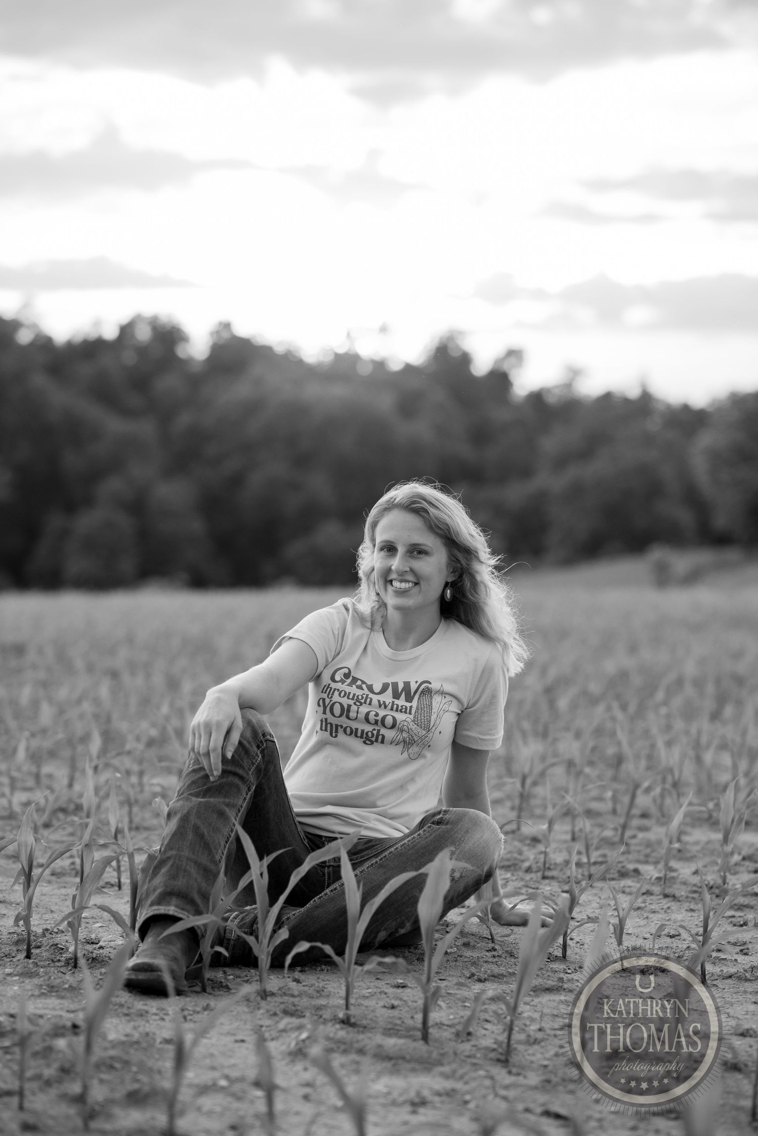 Black and white portrait of someone sitting among young corn stalks in a field.