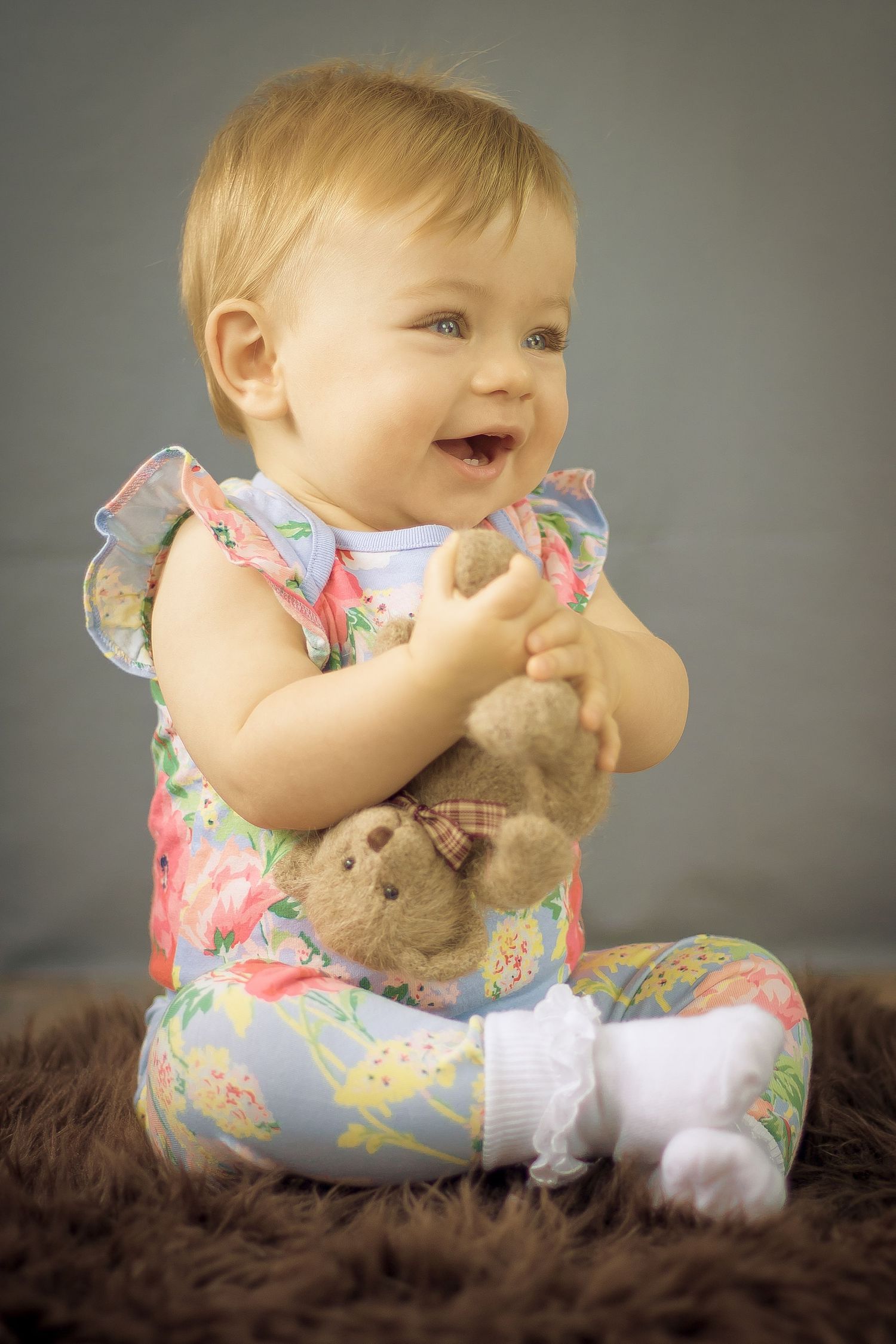 Happy 6 months old holding teddy by the leg in floral jumpsuit.