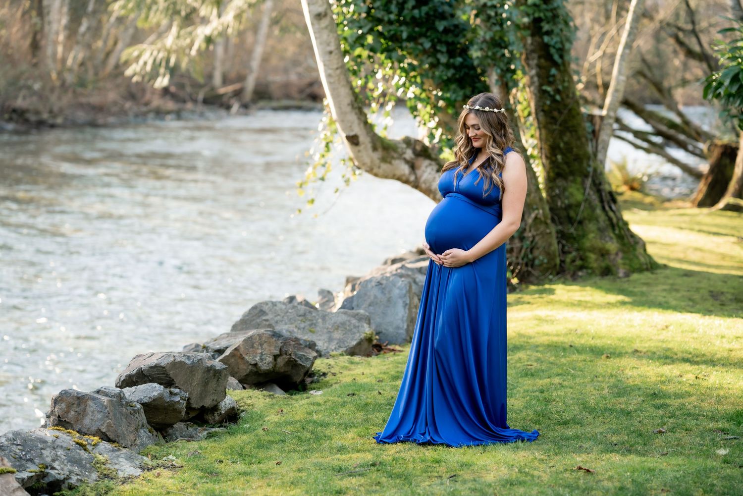 Maternity photo of woman in royal blue dress standing by river with rocks and trees.