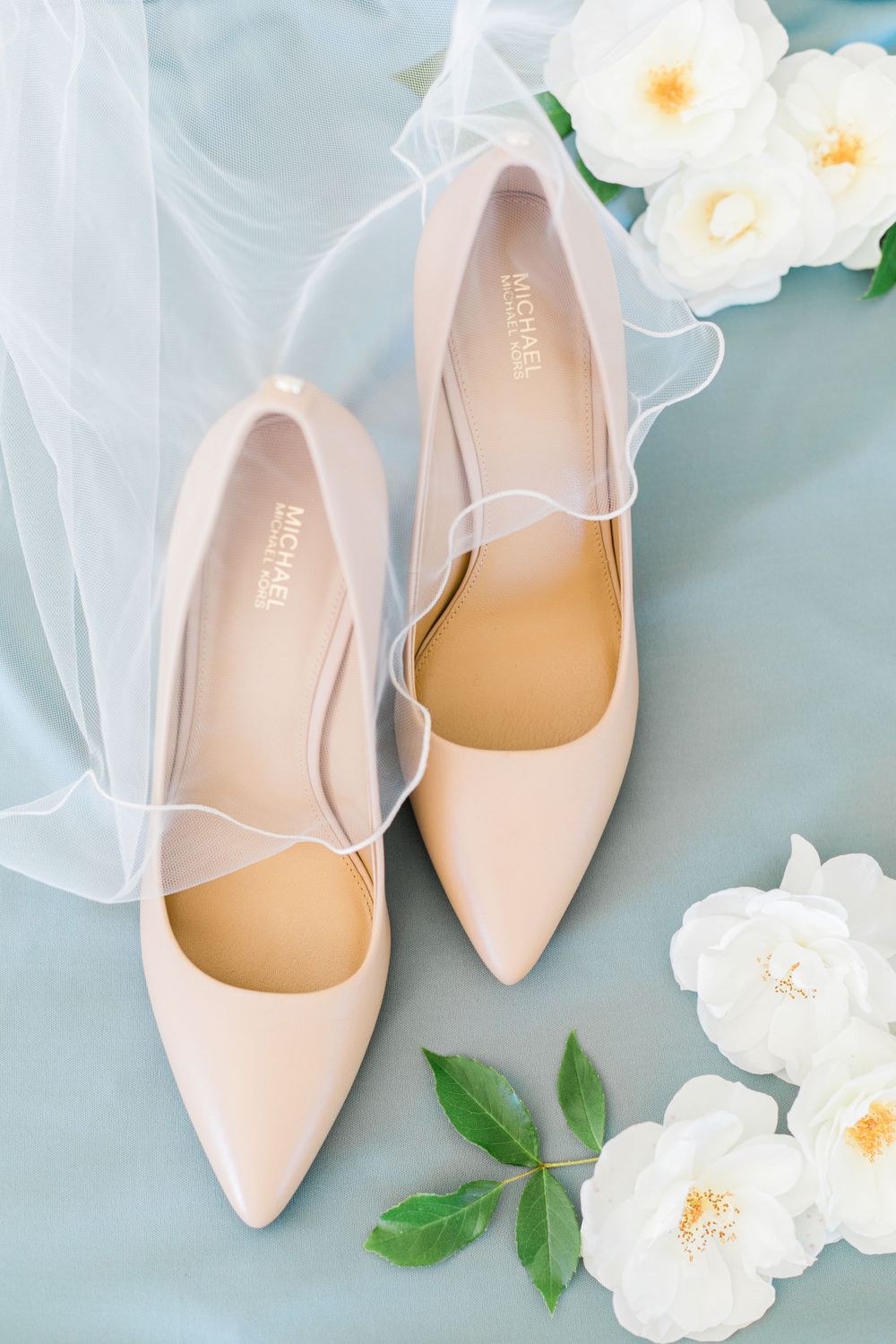 Blush pink pointed ballet flats styled with white roses and sheer veil on blue background.