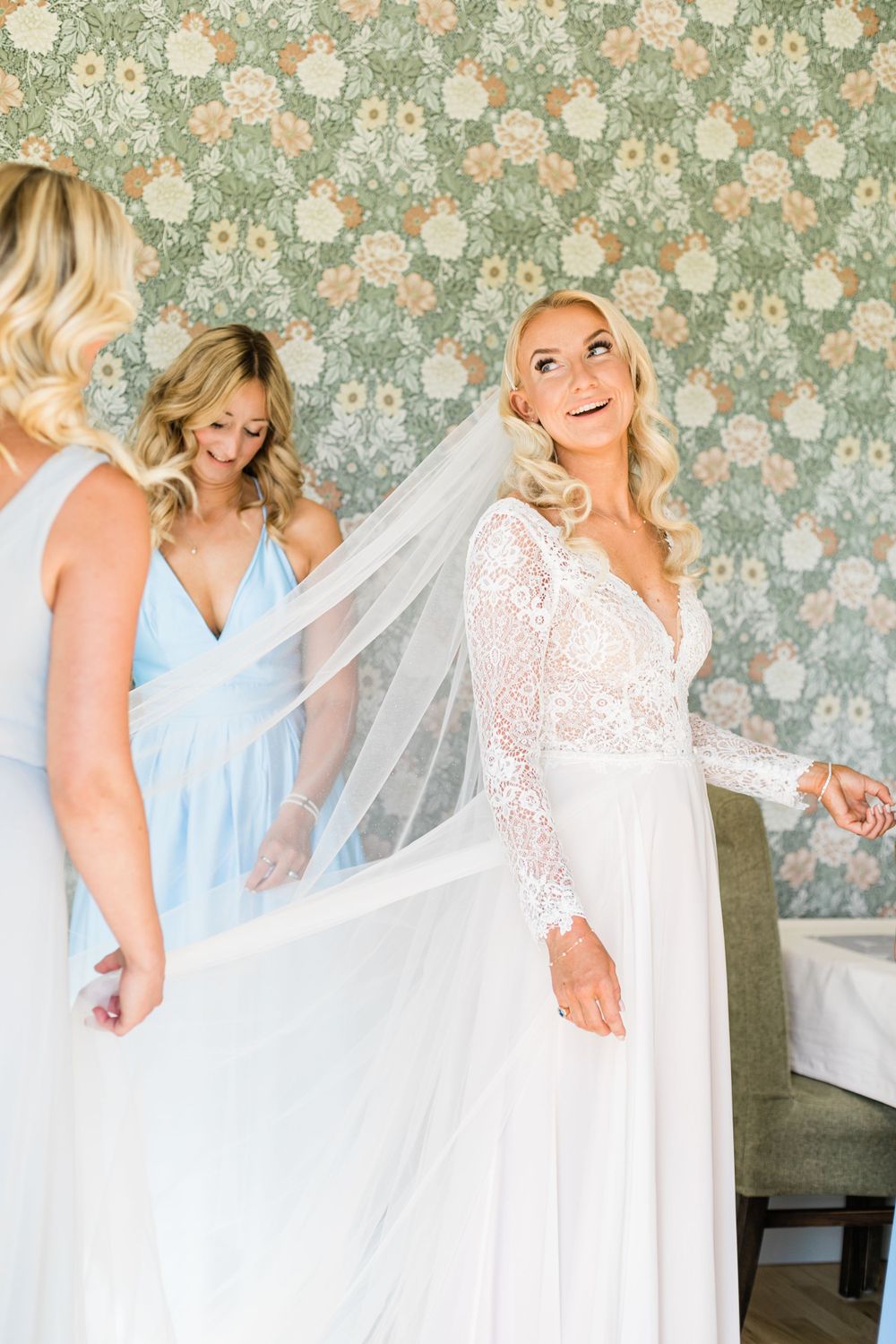 Bride in long sleeve lace wedding gown with bridesmaids in light blue dresses against floral wallpaper.