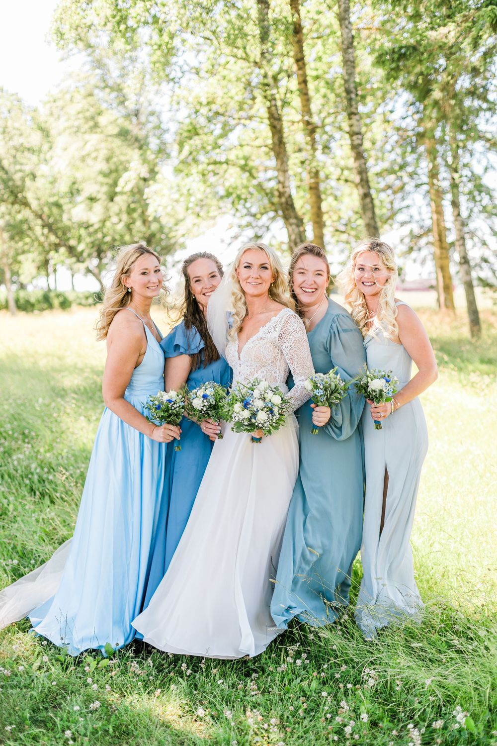 Wedding party in shades of blue dresses pose together in sunny garden setting.