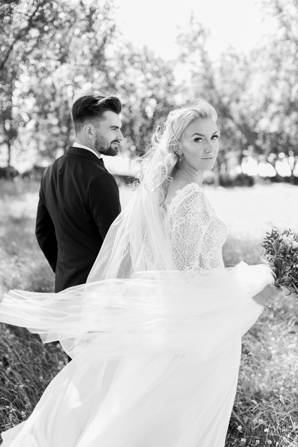 Black and white photo of couple in wedding attire with flowing veil in outdoor setting.