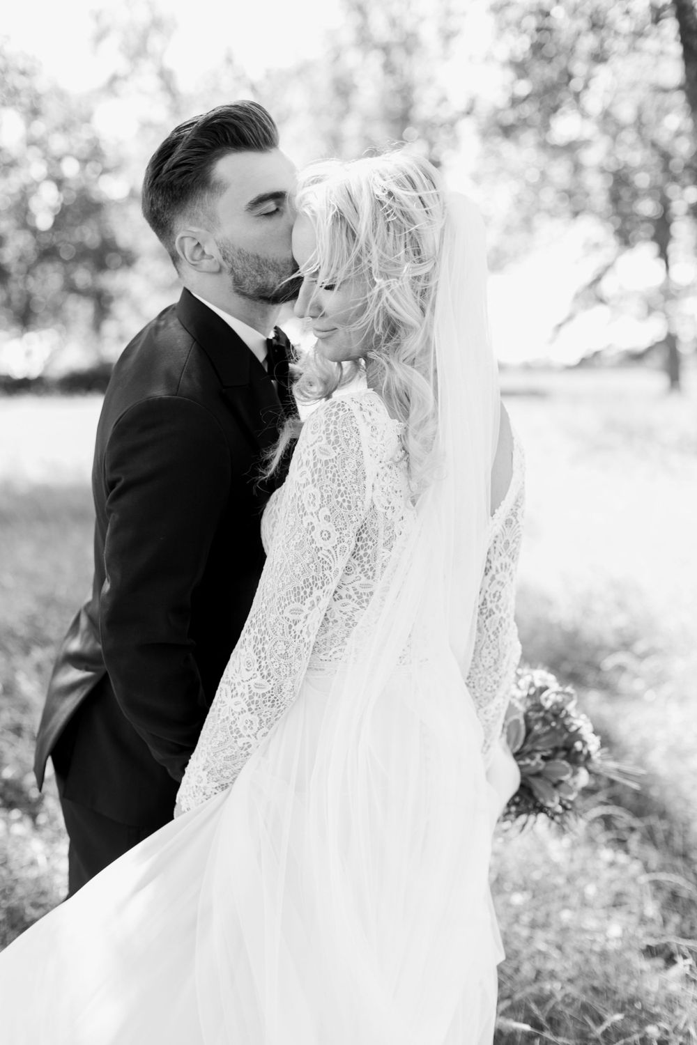 Romantic black and white portrait of newlyweds with flowing veil in natural setting.