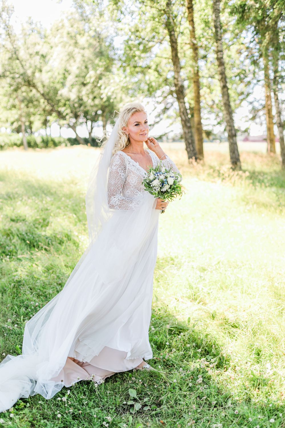 Bride in lace sleeve wedding dress holds bouquet in sun-dappled garden setting.