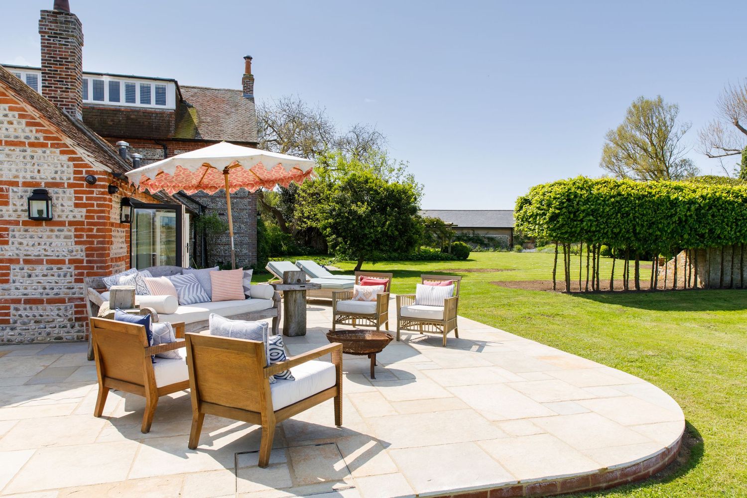 Outdoor patio seating area with umbrella and furniture on stone terrace next to brick house and manicured lawn.