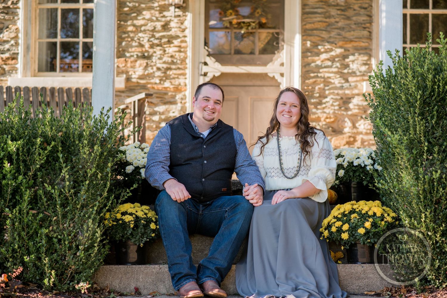 A couple relaxes on stone steps with yellow mums and rustic decor on a stone building porch.
