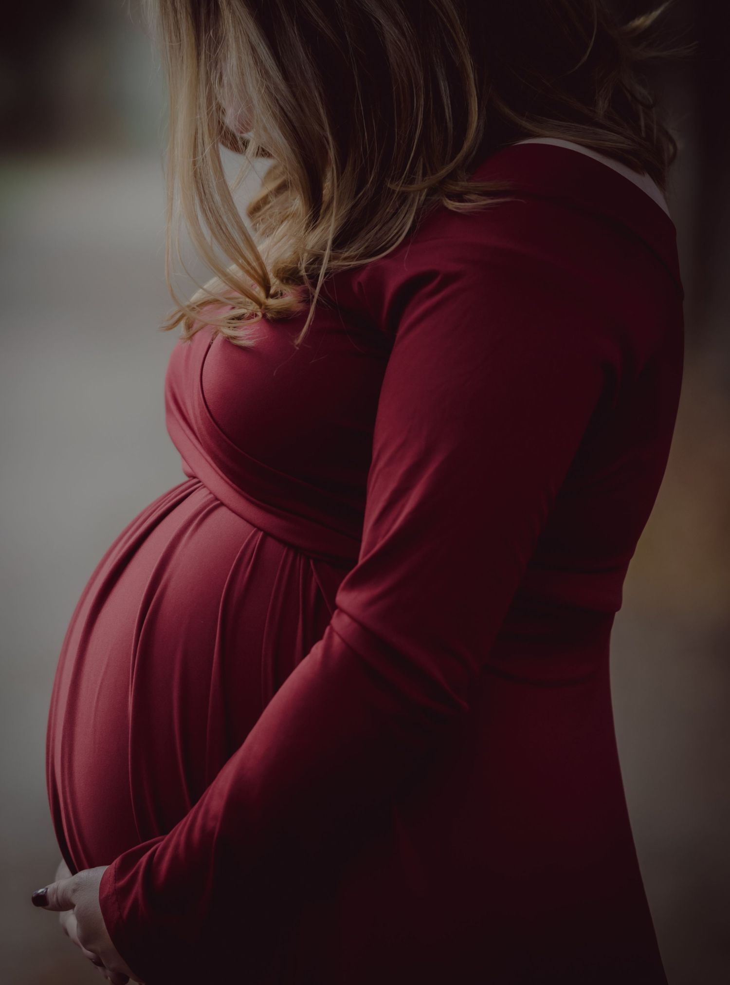 A pregnant woman in a burgundy dress cradles her baby bump against a soft blurred background.