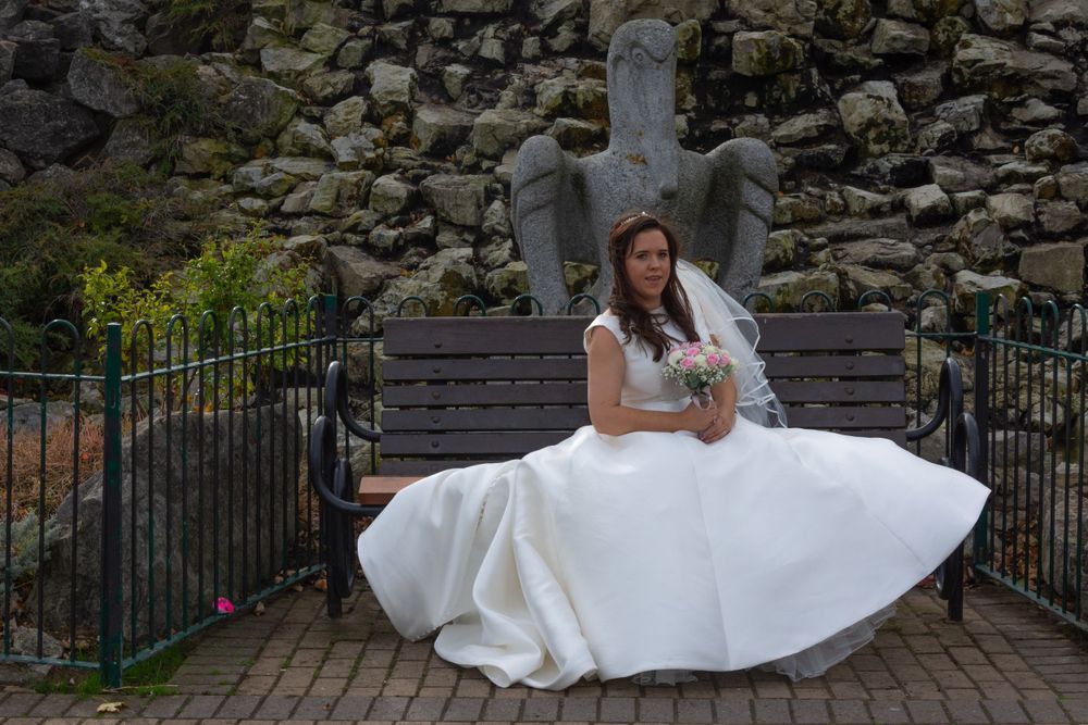 A bride in a white wedding dress sits on a bench with a stone wall and angel statue backdrop.