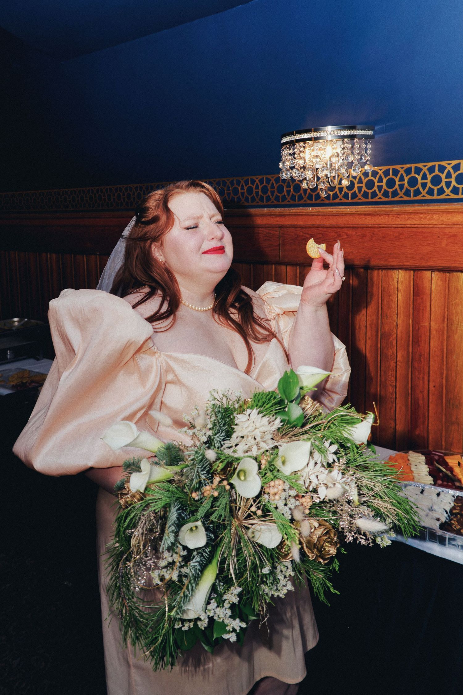 A bride in a champagne satin gown with puff sleeves holds a bouquet and examines a small item indoors.