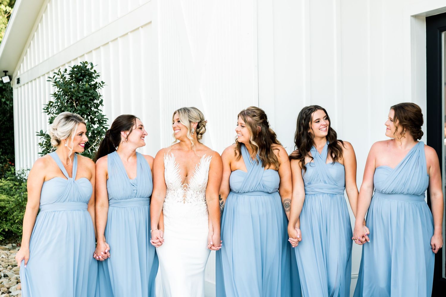 Wedding party members in coordinating blue attire stand in line against white building exterior.