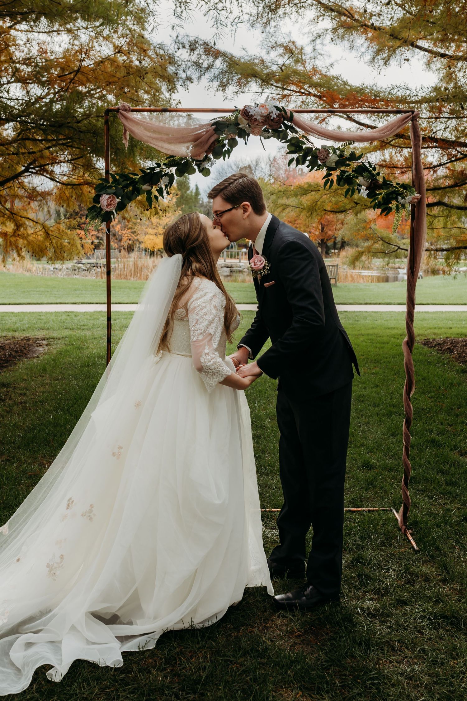 Bride and groom share first kiss under rustic arch decorated with greenery.