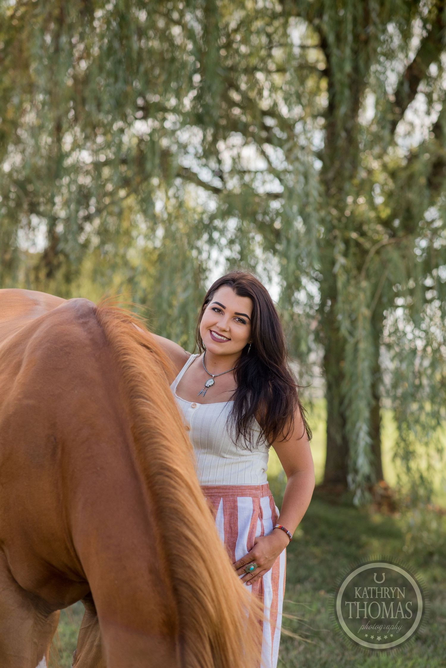 A chestnut horse with flowing mane poses beside someone in a white top under weeping willow trees.