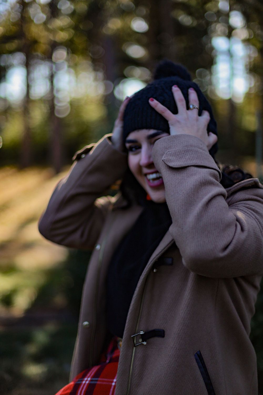 Person in a beige coat and black beanie playfully covers face while laughing outdoors.