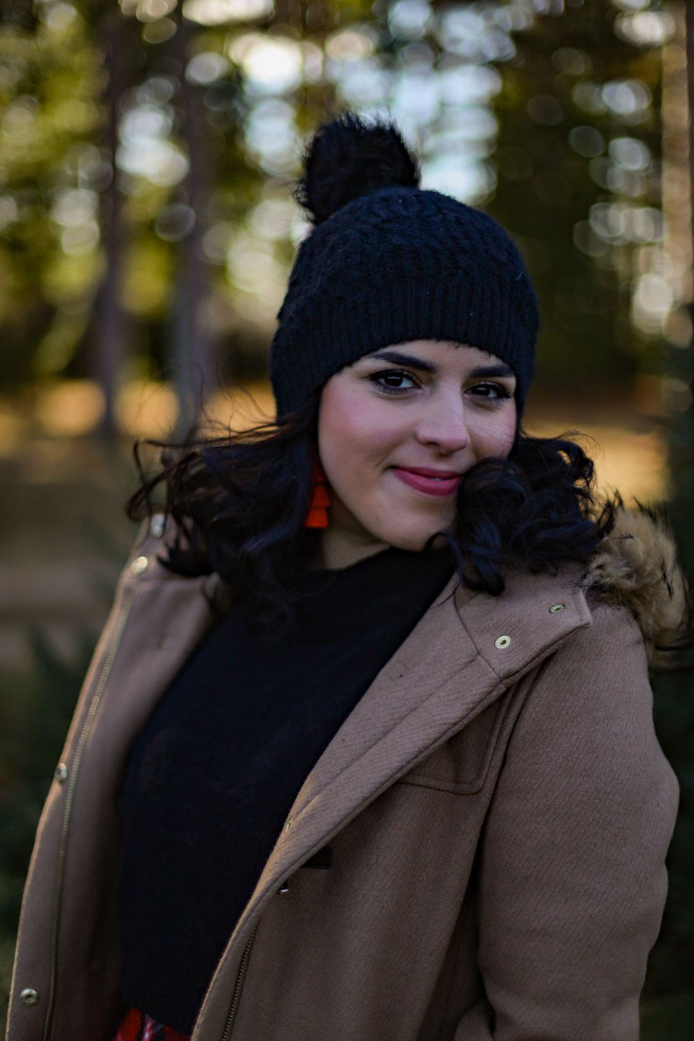 Person in a tan coat and black beanie hat smiles at the camera on a winter day.