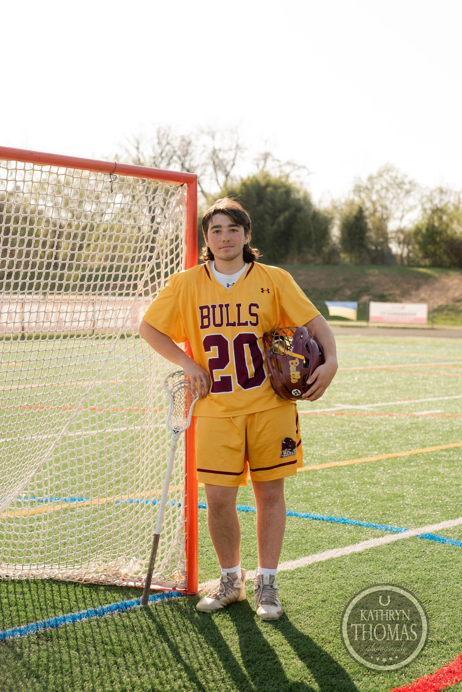 Athlete in yellow Bulls uniform number 20 standing with lacrosse gear by goal net on field.