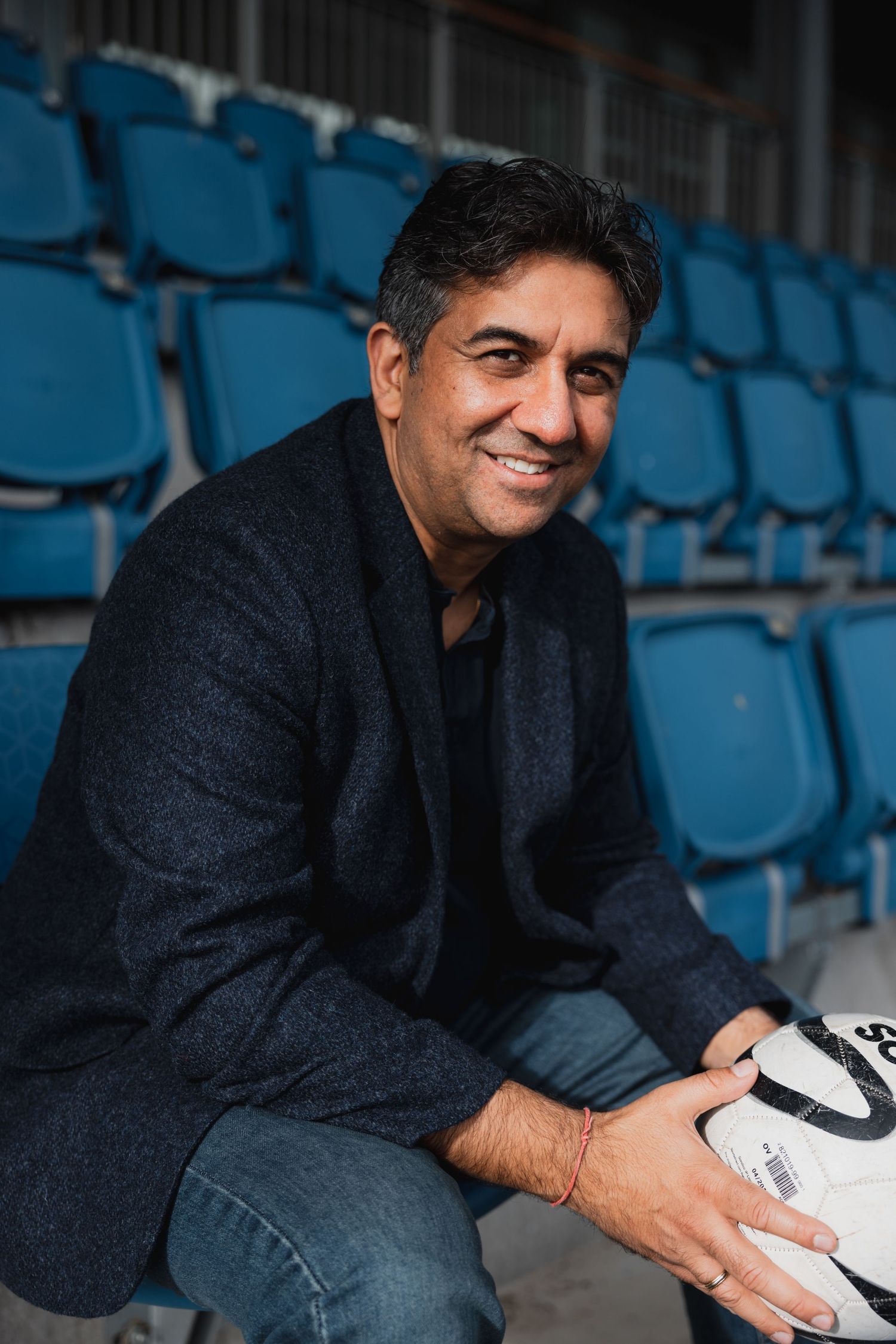 Indian man sitting in a football stand wearing a blazer and jeans and holding a football