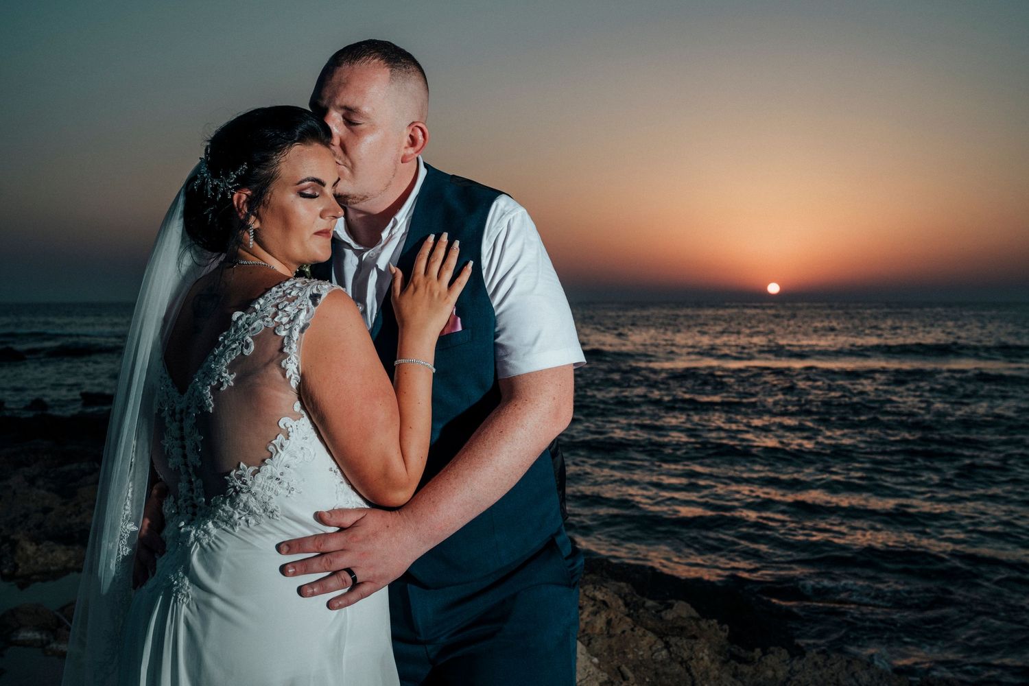 Romantic sunset wedding portrait of a couple embracing on a beach with orange sky and ocean backdrop.