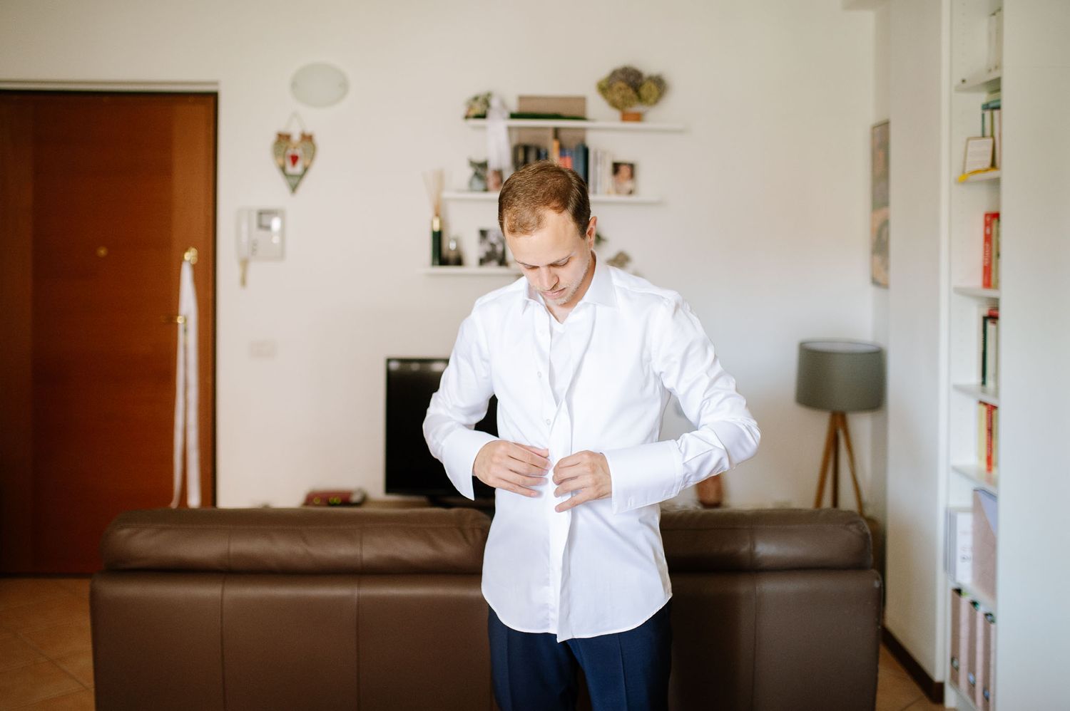 Well-dressed man in white shirt buttoning up his collar in a living room with brown leather sofa.