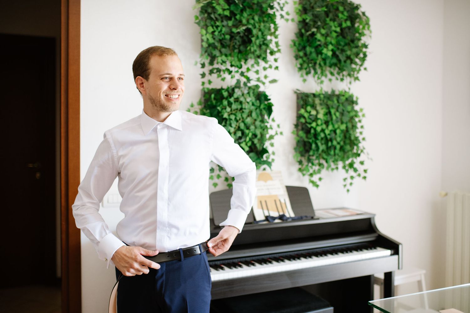 Man in white shirt stands next to black piano with hanging plants in background.