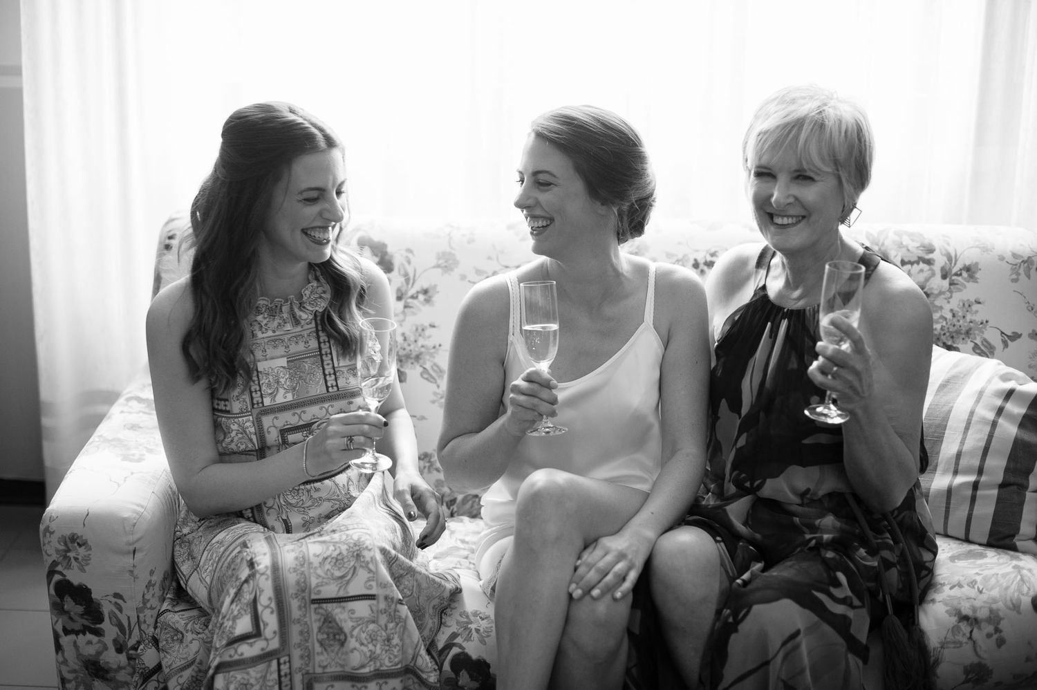 Black and white photo of three friends sharing champagne and laughter on a couch.