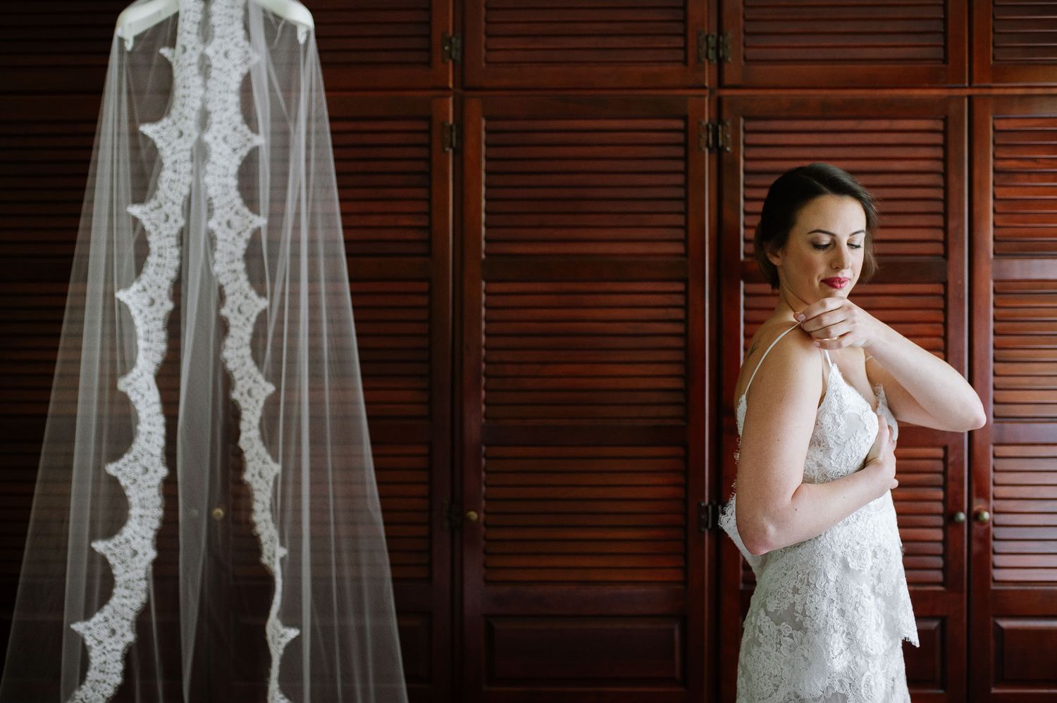 A wedding dress and veil hanging beside wood shutters as someone adjusts their jewelry.