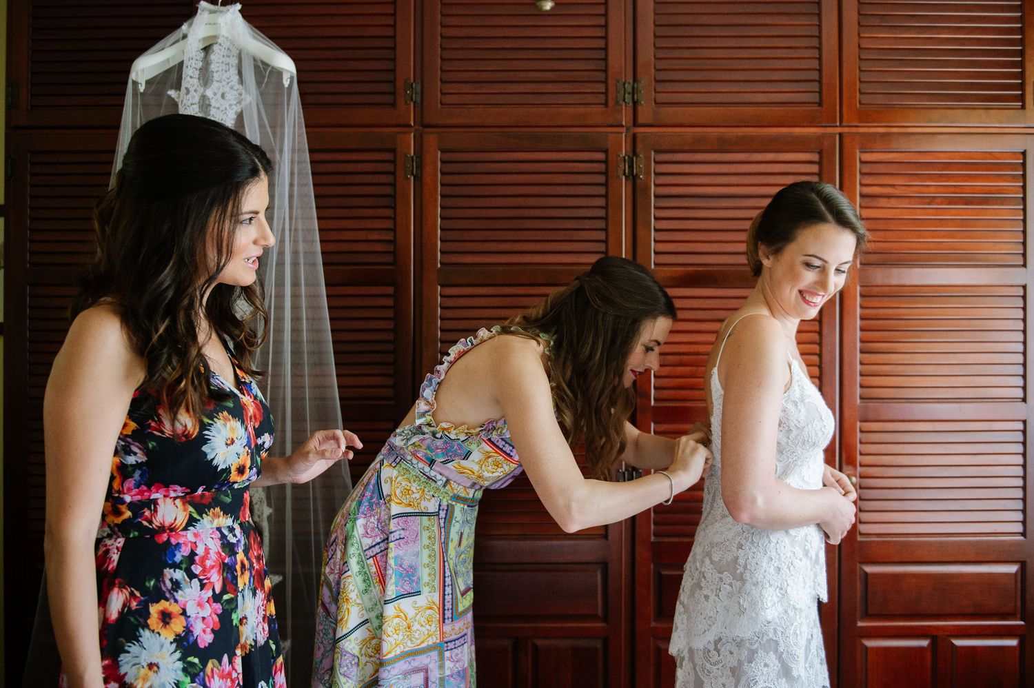 Women helping with wedding dress preparations in front of wooden closet doors.