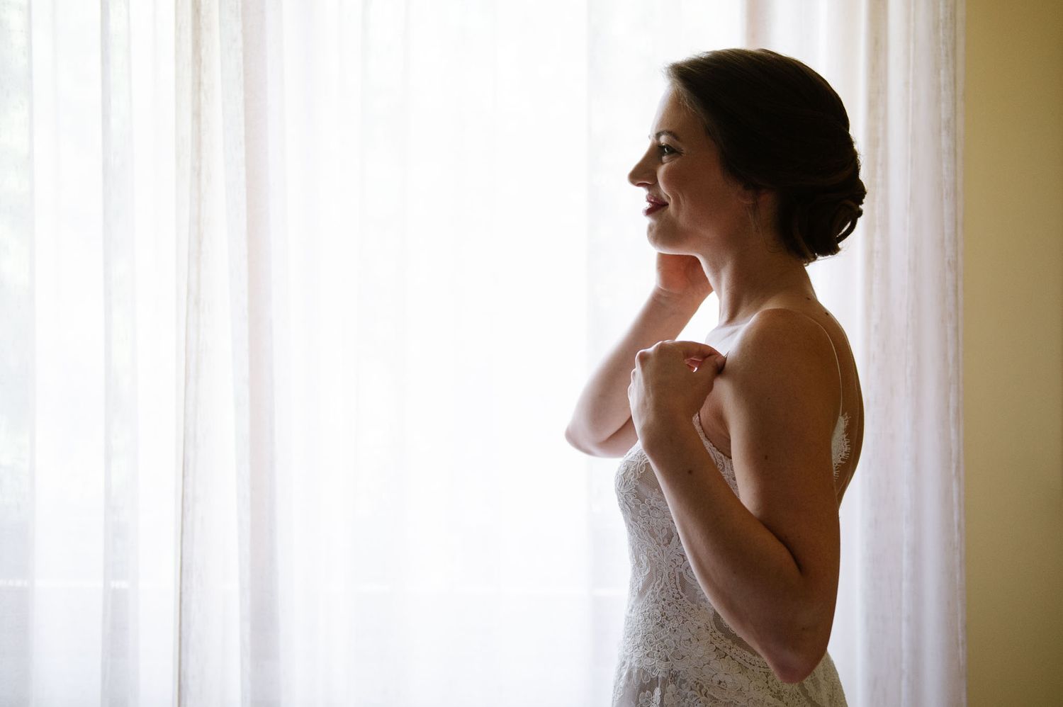 Silhouette of person in wedding dress against bright window adjusting jewelry.