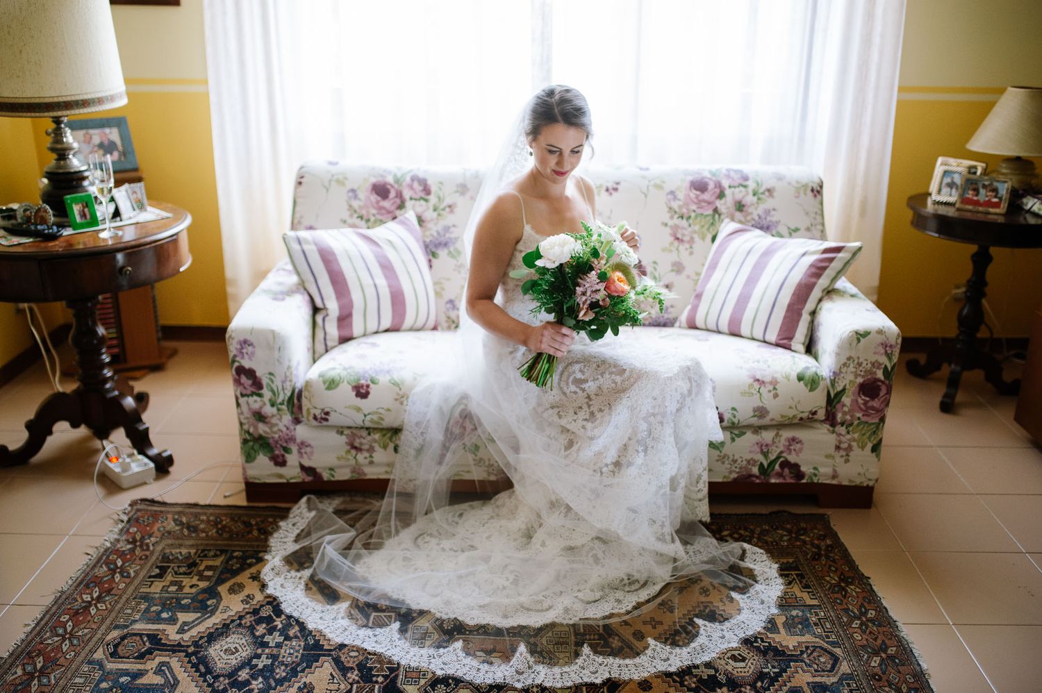 Wedding dress billowing over floral couch as bouquet rests in lap.