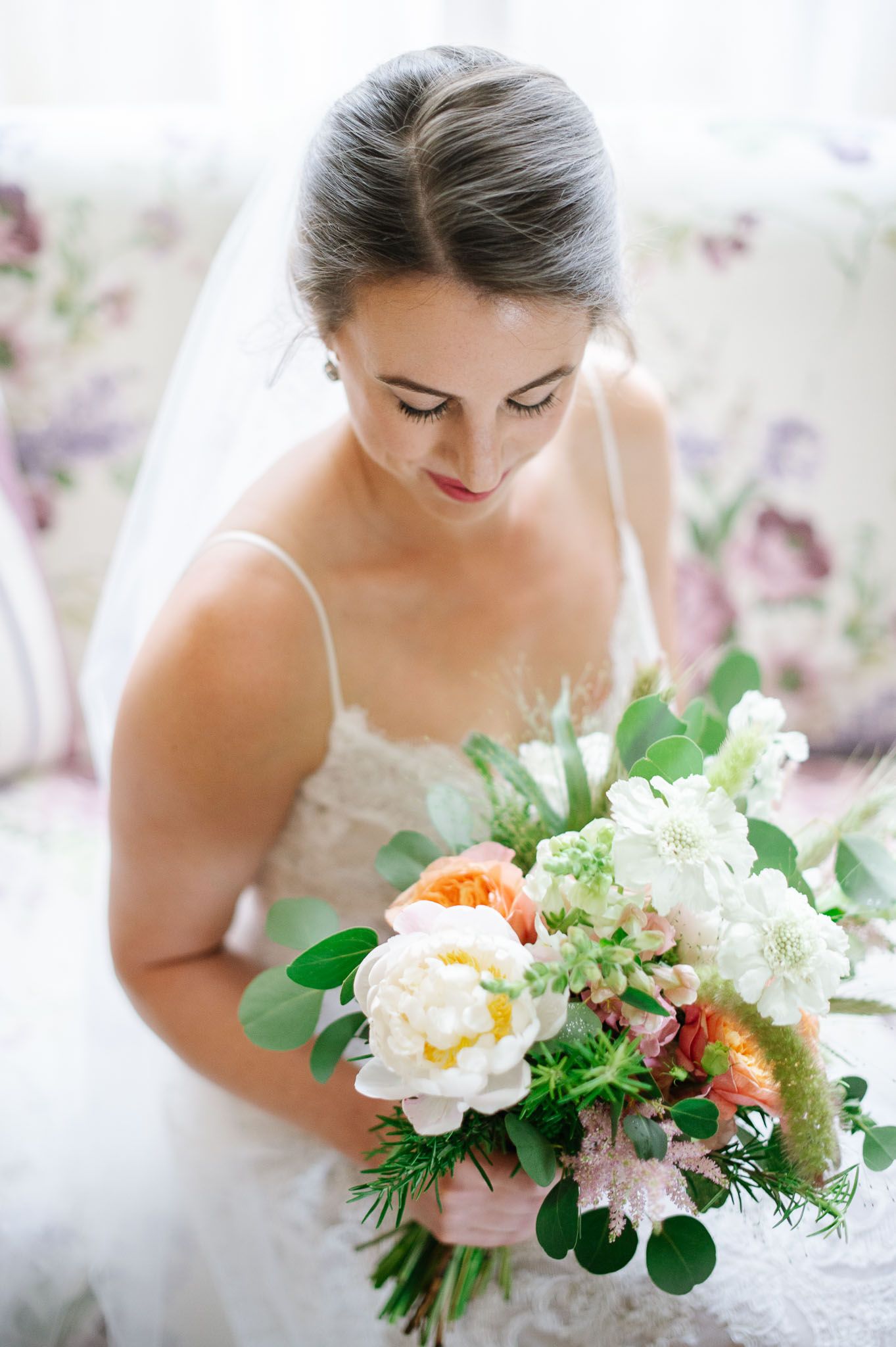 A delicate wedding bouquet with white and peach peonies is held by a bride in a lace dress.