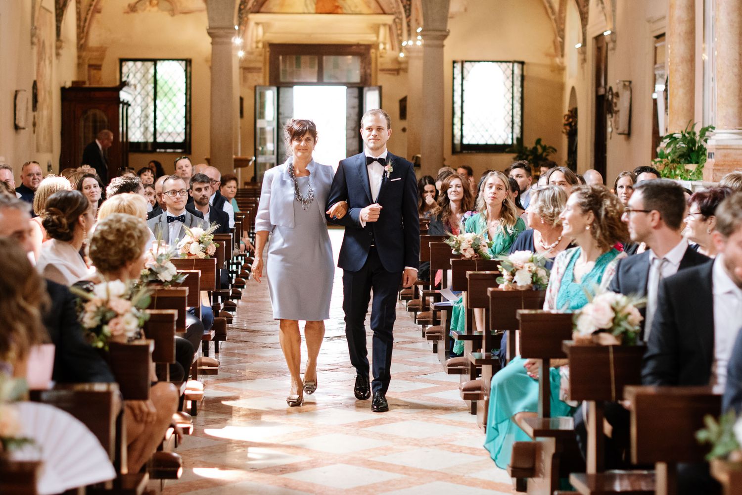 Guests seated in church pews during wedding ceremony with decorative floral arrangements.
