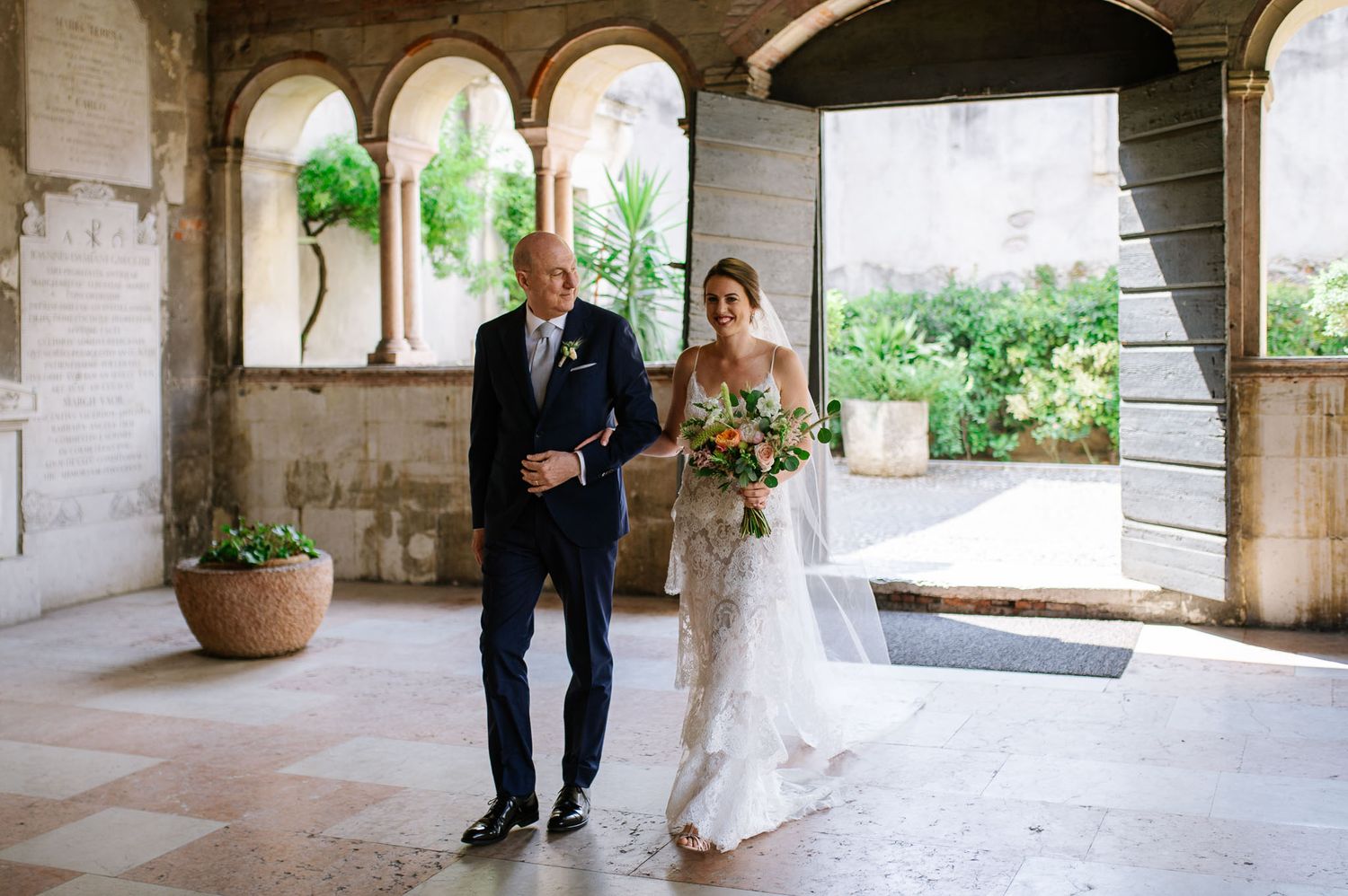 Elegant covered walkway with stone arches and potted plants during wedding ceremony.