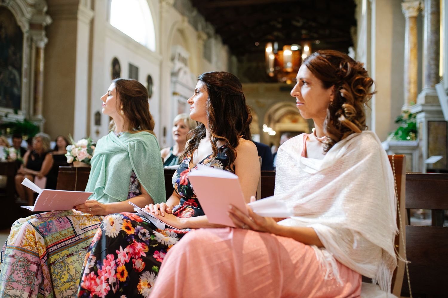 Three elegantly dressed attendees sit in ornate church pews during a wedding ceremony.