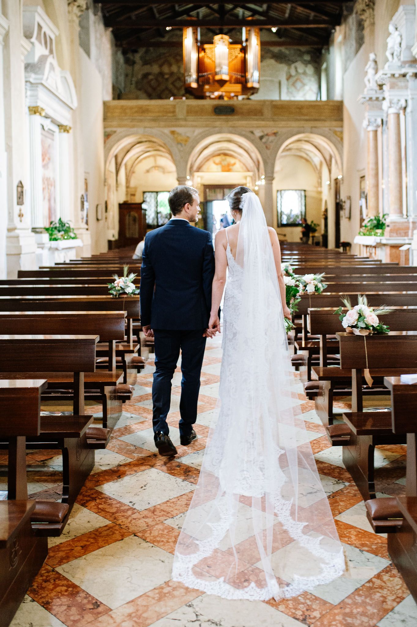 A couple walks down the aisle of an elegant church with a flowing veil trailing behind.