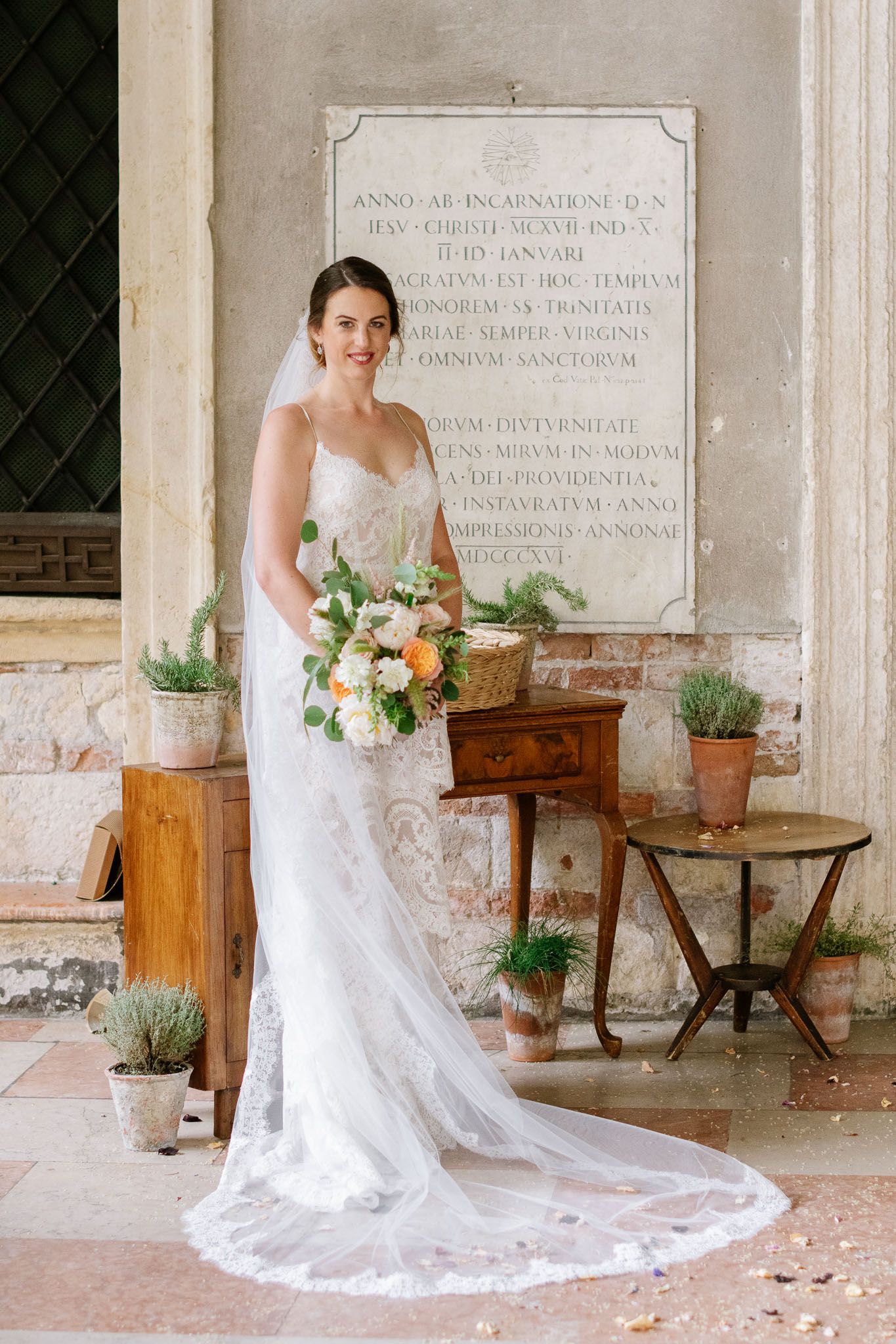 A bridal portrait in front of a stone wall with potted plants and elegant lace wedding dress with long train.