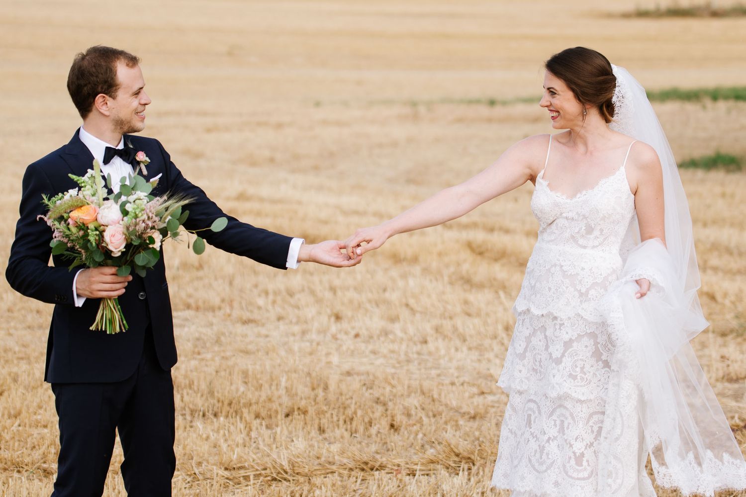 A couple holds hands while walking through a golden wheat field during their wedding photoshoot.