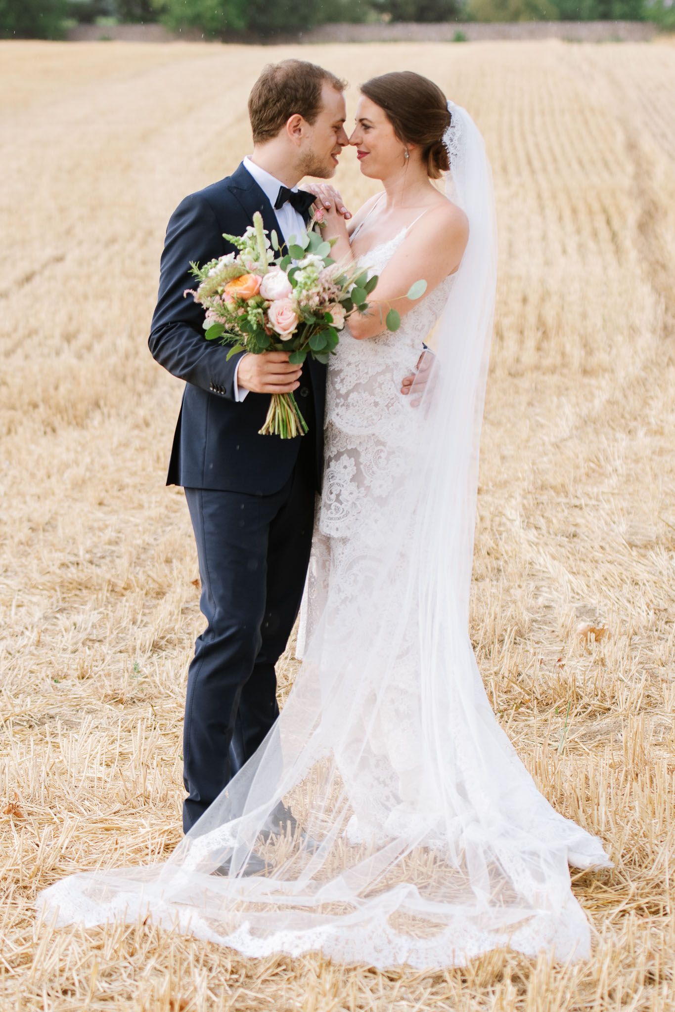 A romantic moment between newlyweds standing in a wheat field with flowing veil and floral bouquet.