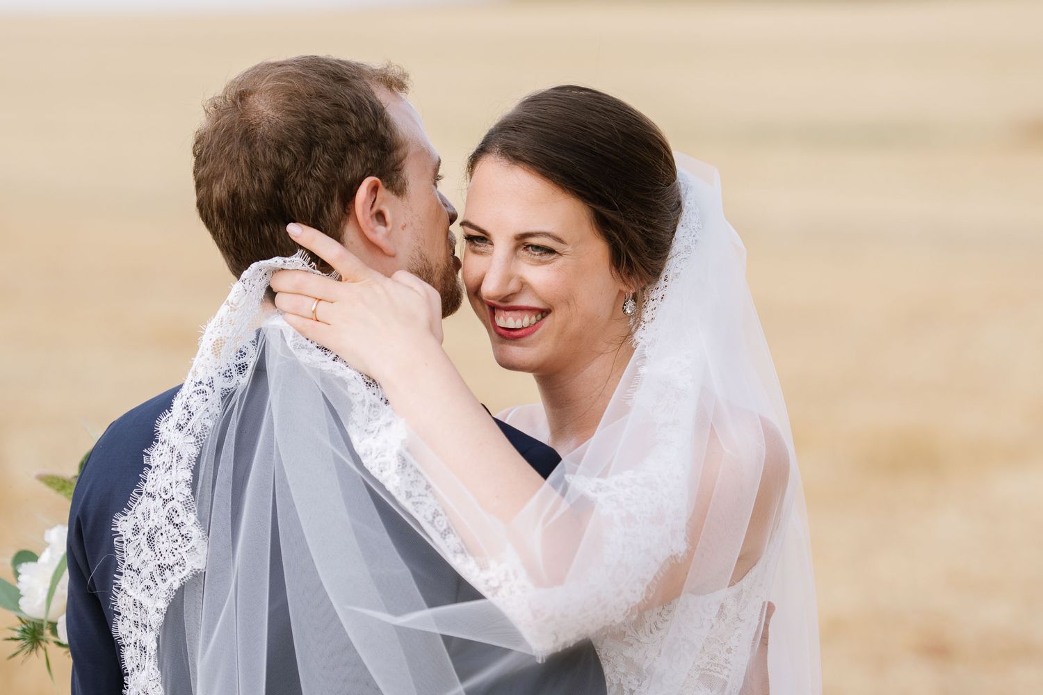 A tender embrace between newlyweds with delicate lace veil against a wheat field backdrop.