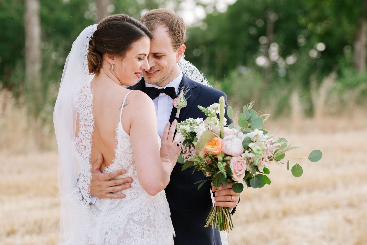 An intimate wedding portrait with couple embracing among trees while holding a colorful bouquet.