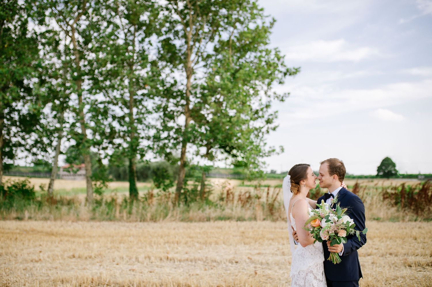 A romantic wedding couple embraces in a sun-drenched field with green trees in the background.