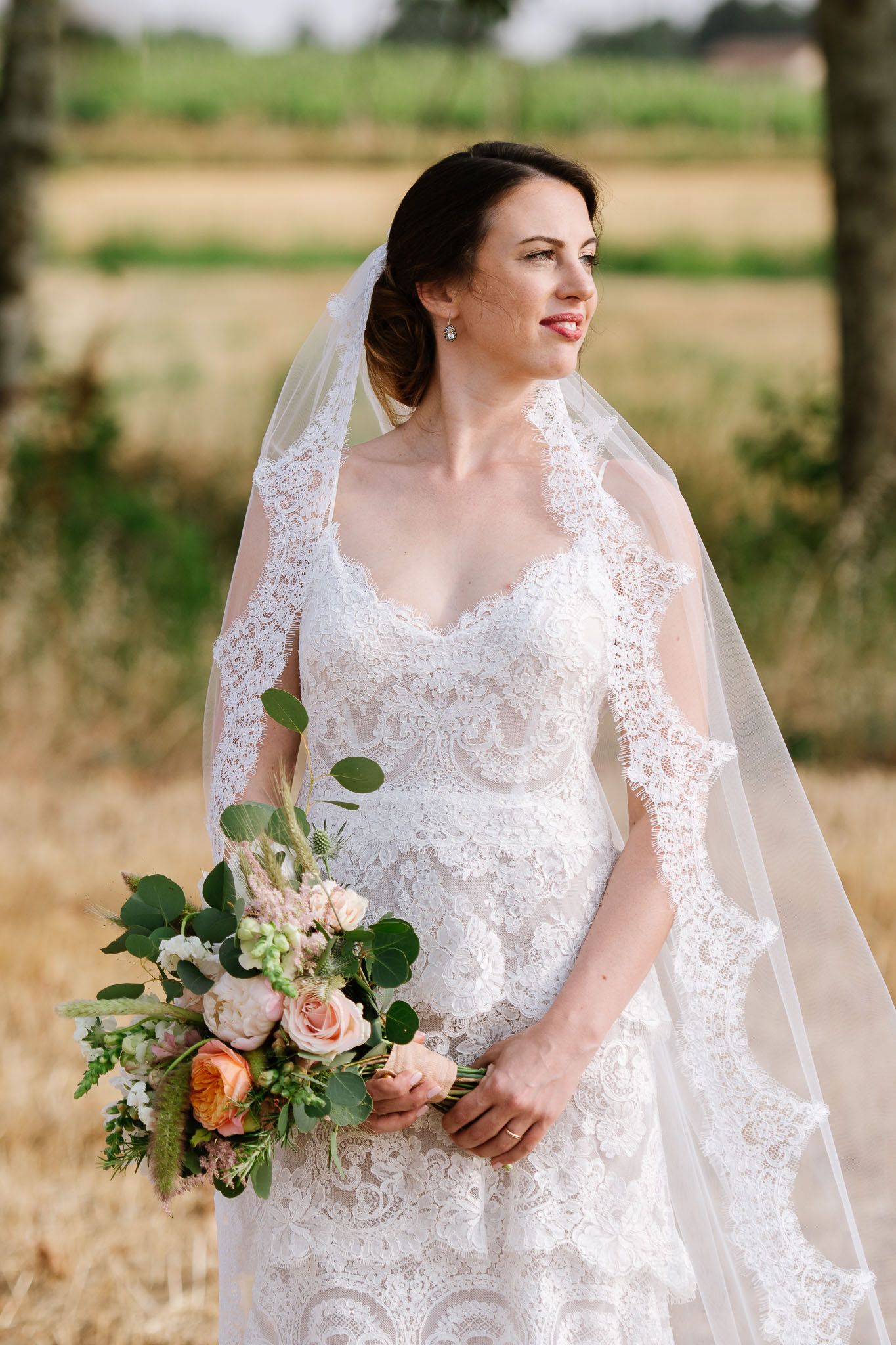 A bride wearing a detailed lace wedding dress and veil holds a pink and green floral bouquet.