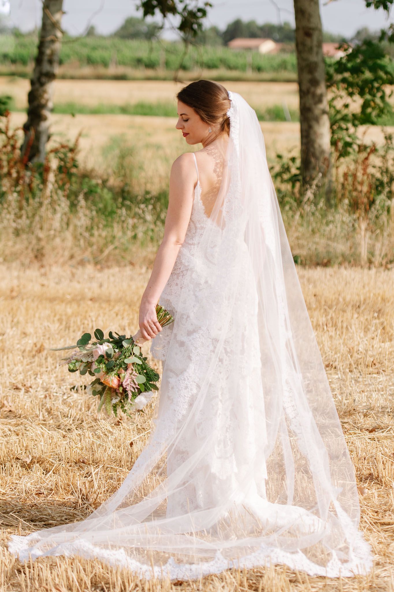 A bride in a lace wedding gown with flowing veil poses in a rustic field holding a floral bouquet.