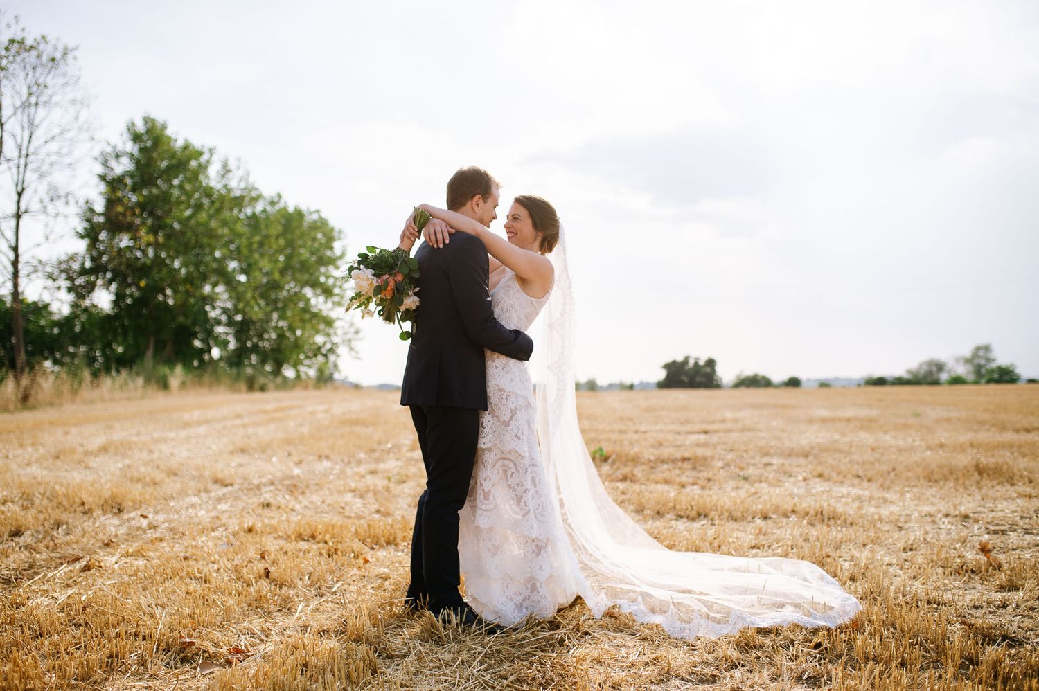 Newlyweds share an intimate moment in a sunlit field with trailing white veil.