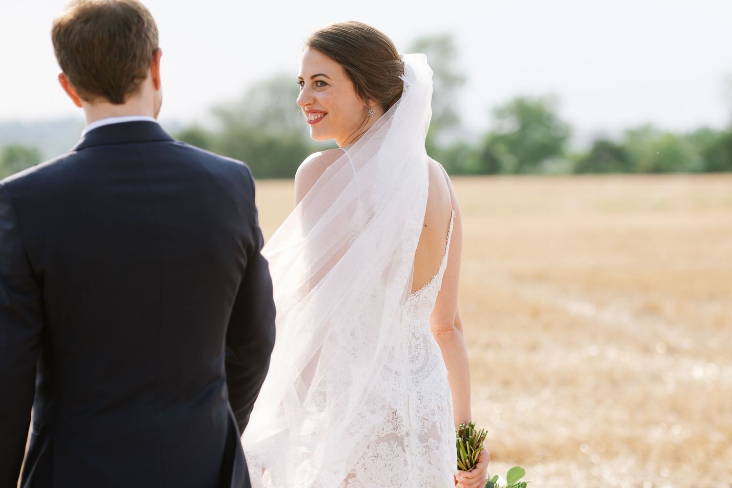 A wedding couple stands together in formal attire against a golden field backdrop.