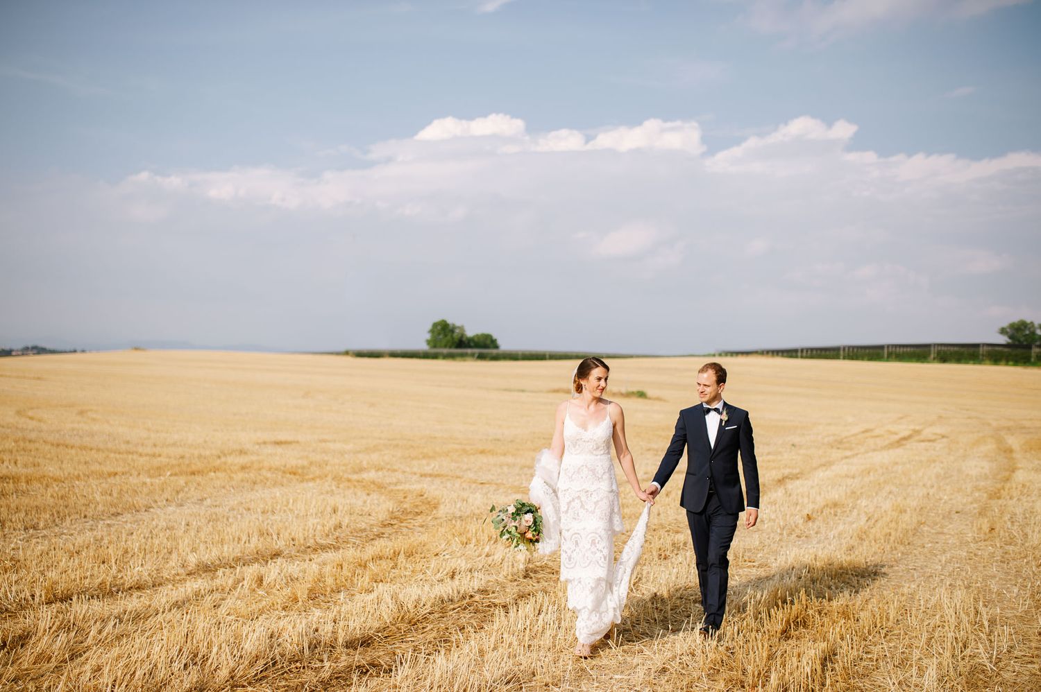 A wedding couple walks hand in hand through a golden wheat field under blue skies.