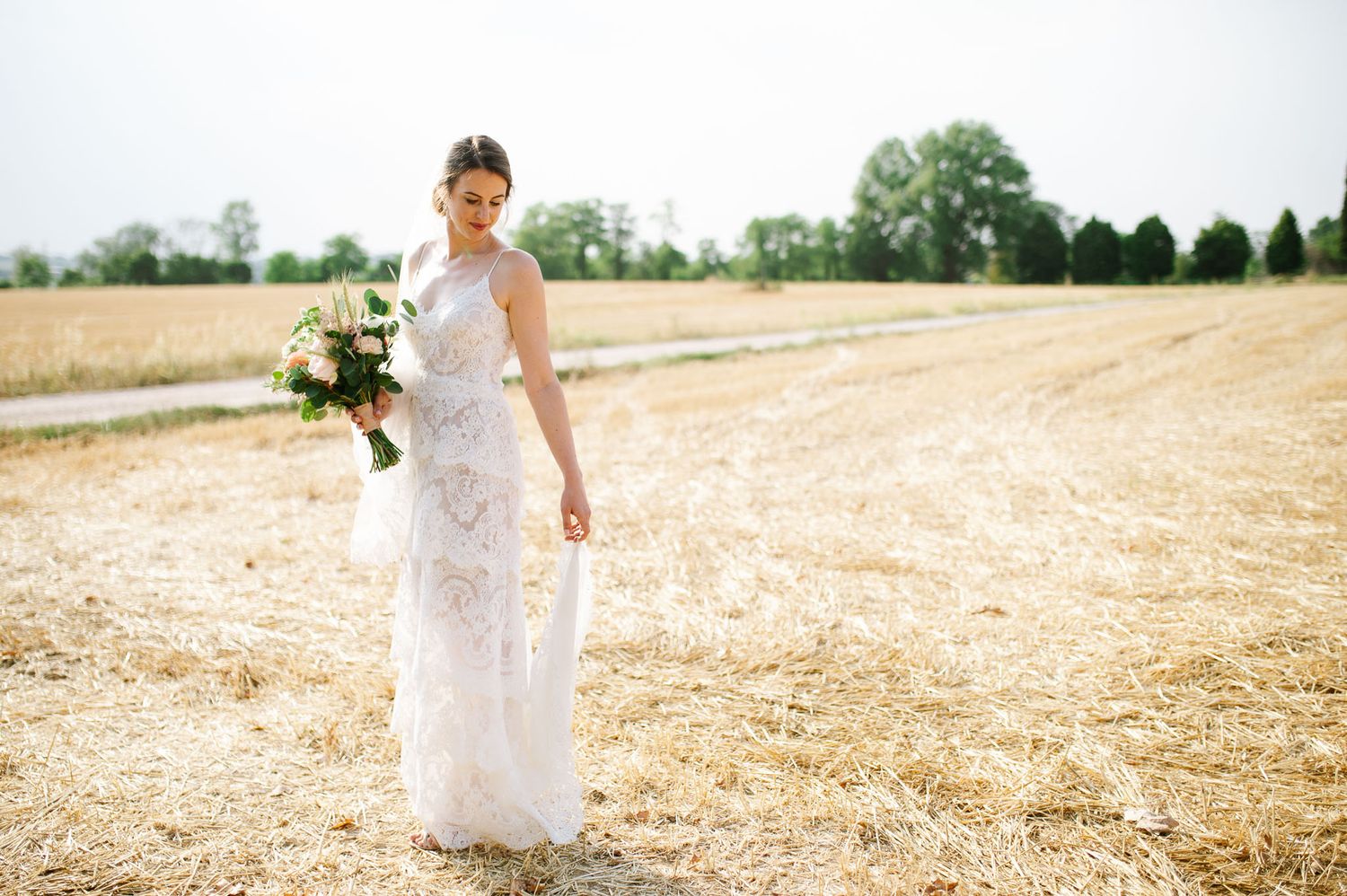 A bride in lace gown poses in sunlit field holding a fresh floral bouquet.